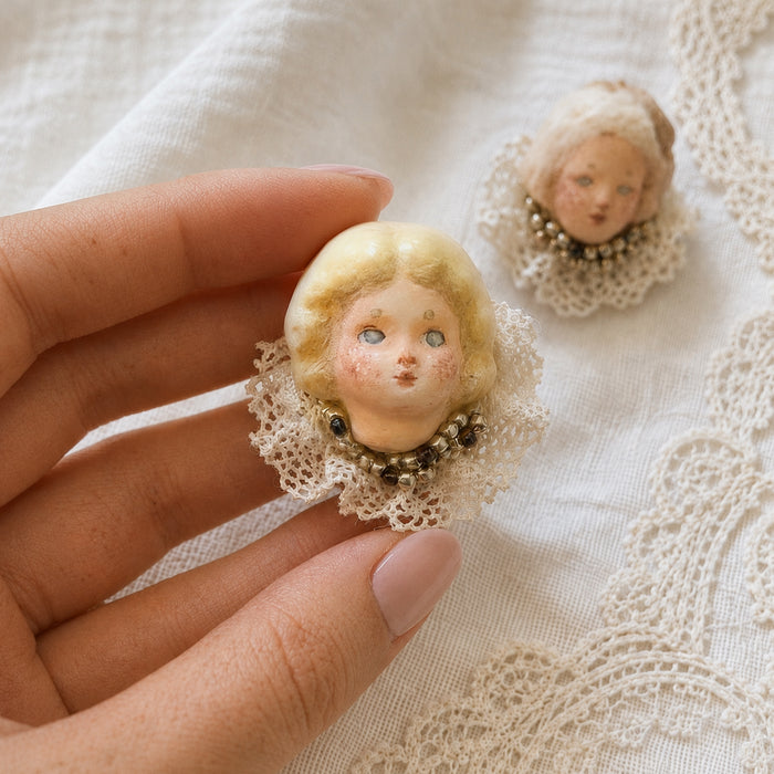 Lambwick doll ring with blonde curls, rosy cheeks and a cream lace collar, held between fingers.